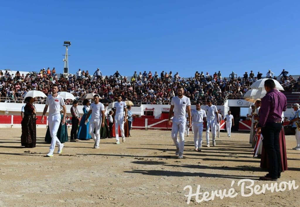 Procession d'hommes en blanc et femmes en costumes traditionnels dans une arène bondée sous le soleil, foule dans les gradins. TORI.
